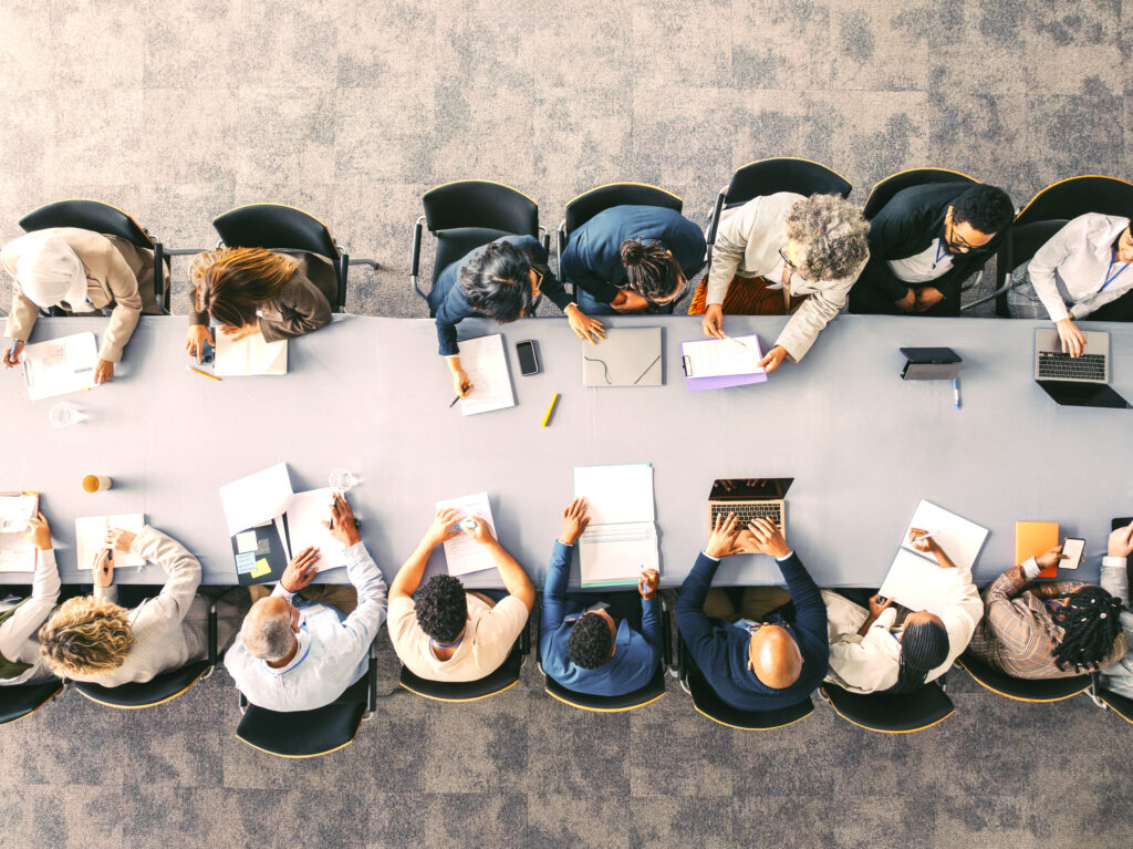 Das Bild zeigt eine Gruppe von Personen an einem langen Tisch, die miteinander interagieren.