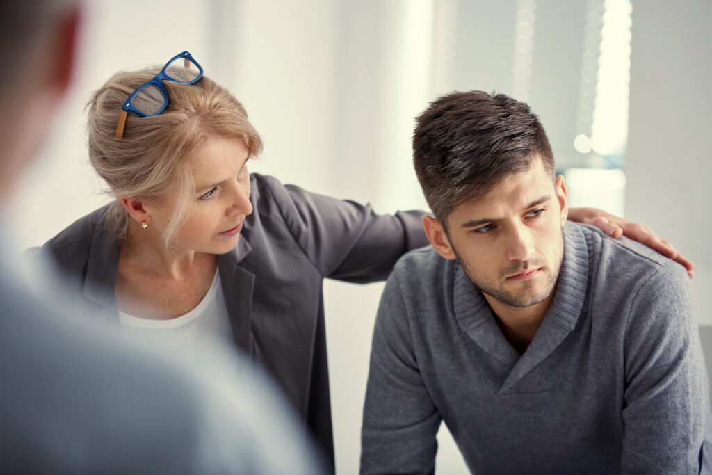 Das Bild zeigt 3 Personen, wovon die Frau in der Mitte einen jungen Mann tröstet, welcher sich gerade bei seinem Therapeuten befindet.